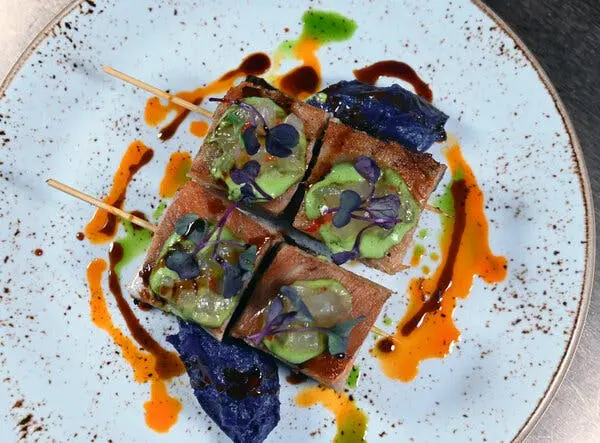 نمای بالای چهار تکه مربعی غذا روی سیخ‌ها، که با آووکادوی سبز و میکروگروه‌های بنفش پوشیده شده‌اند، روی بشقاب نقطه‌دار با قطره‌های سس رنگارنگ.