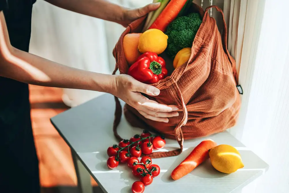 تصویر برشخورده از زن آسیایی جوان که میوهها و سبزیجات را از کیف خرید سازگار با محیطزیست در میآورد.
